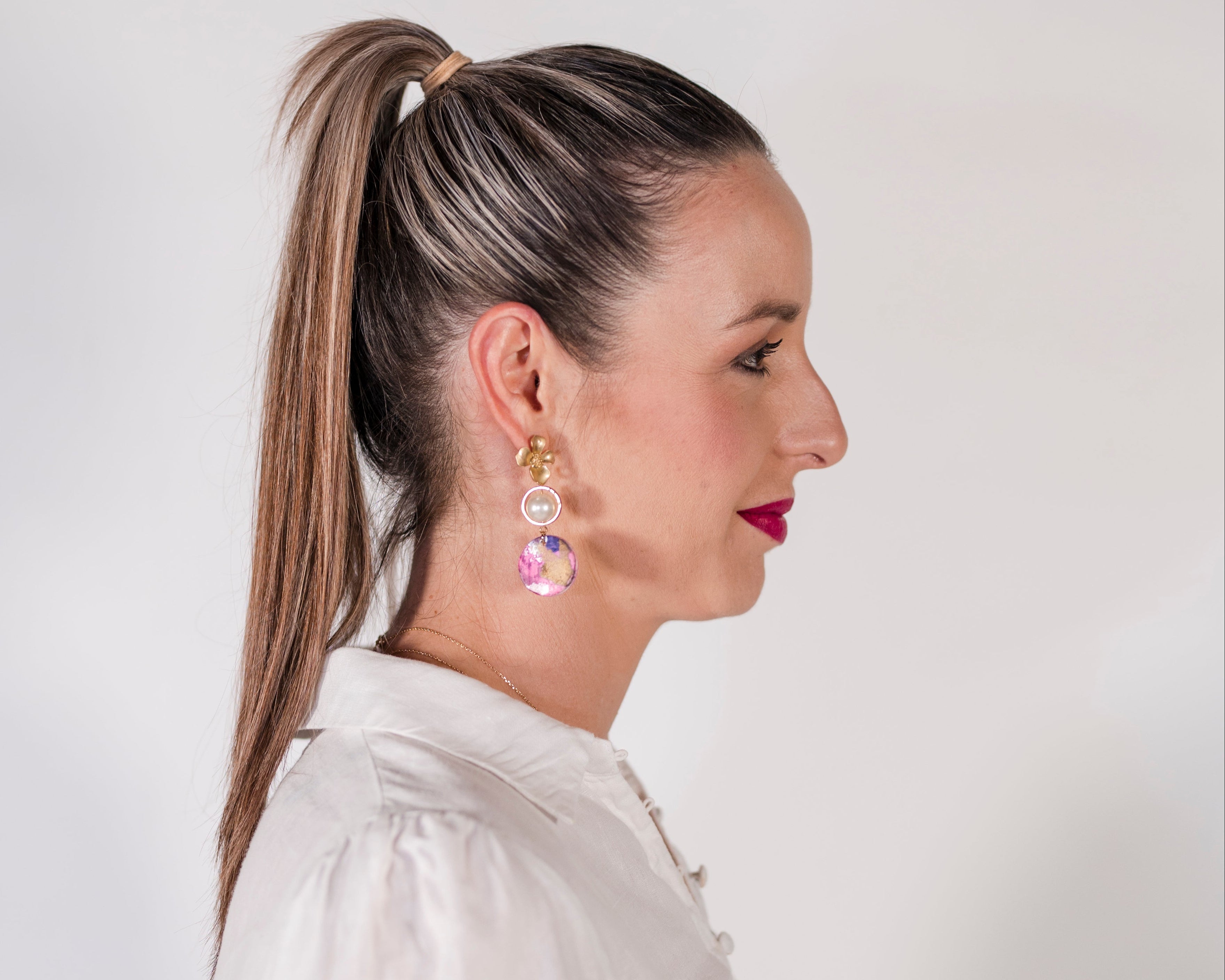 Woman wearing a white shirt with a ponytail and colorful earrings on a plain background